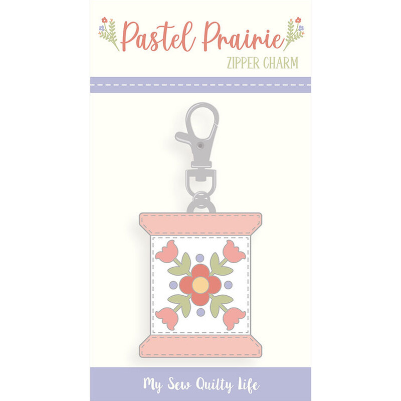 An enamel and metal zipper pull in its packaging in the shape of a thread spool with a floral design from the Pastel Prairie fabric collection.