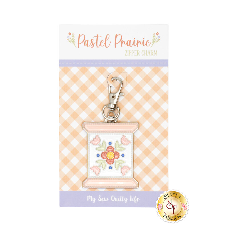 An enamel and metal zipper pull in its packaging in the shape of a thread spool with a floral design from the Pastel Prairie fabric collection.