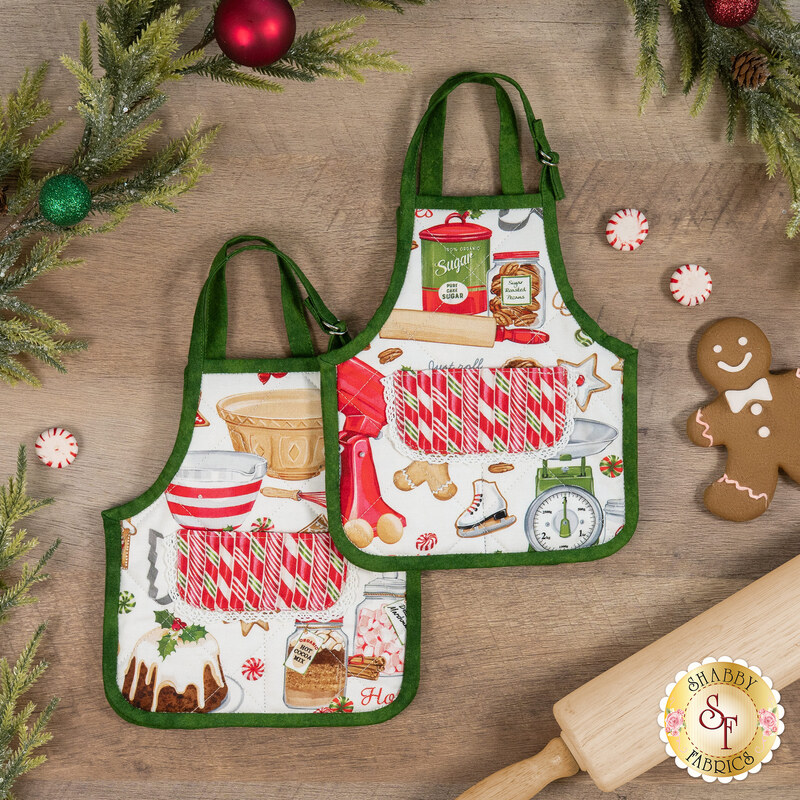 Two festive aprons are staged on a rustic wooden table, decorated with holiday baking motifs and greenery.