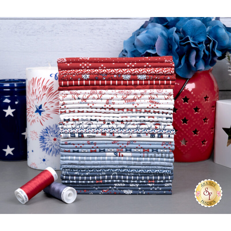 A stack of the fabrics included in the Independence Day FQ set, with jars and flowers and a candle and thread.