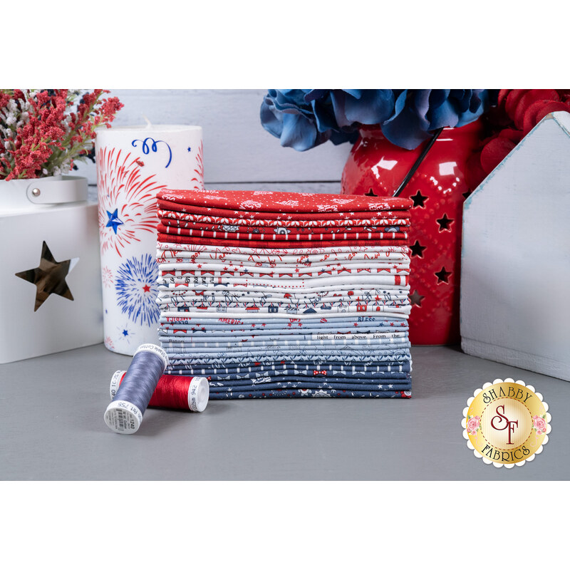 A stack of fabrics included in the Independence Day F8 set, with a jar and a candle and flowers and thread.