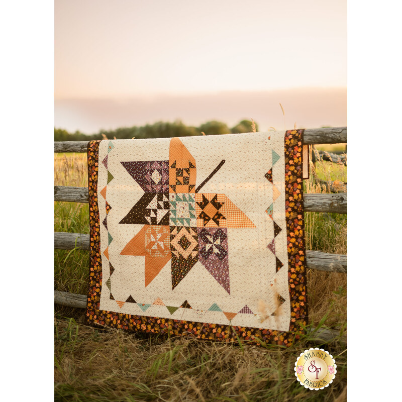 The completed Pressed Leaves quilt colored in autumn fabrics from the The Awe of Autumn collection, hung over a wooden fencepost in front of a field at sunset for a rustic look.