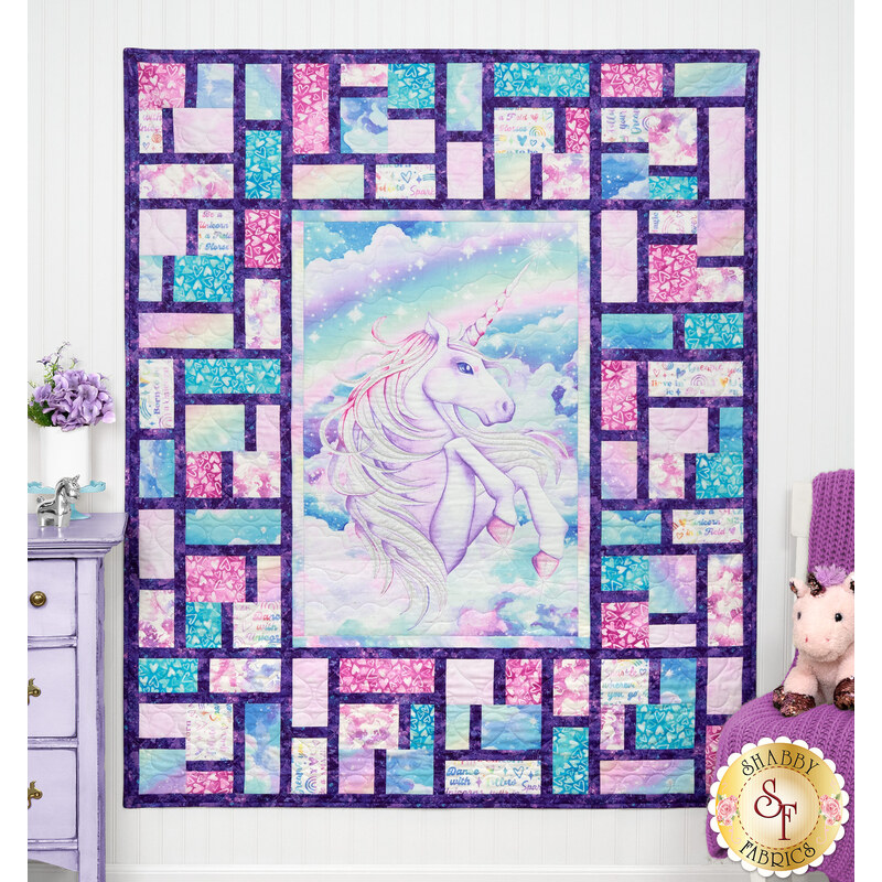 The completed Stained Glass Panel Quilt, colored in blue, pink, and white fabrics from the Sparkle Magic collection by Northcott Fabrics, hung on a lilac paneled wall and staged with coordinating unicorn decor.