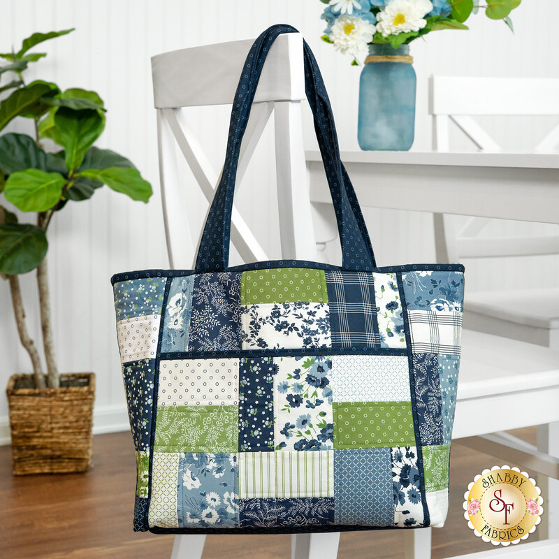 The completed Charming 8 Pocket Tote, colored in navy, blue, white, and green fabrics from the Nantucket Summer collection, hung on the back of a white chair at a table staged with coordinating floral décor.