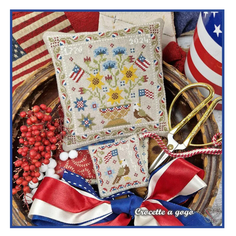 A image of the completed Patriotic Basket cross stitch pattern staged with American flags, ribbon and scissors..