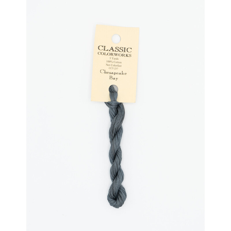 A skein of blue gray embroidery floss, twisted and bundled, with a cream and black label featuring the brand name and color information.