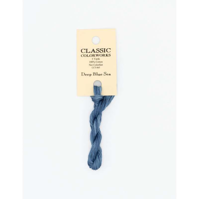A skein of medium blue embroidery floss, twisted and bundled, with a cream and black label featuring the brand name and color information.