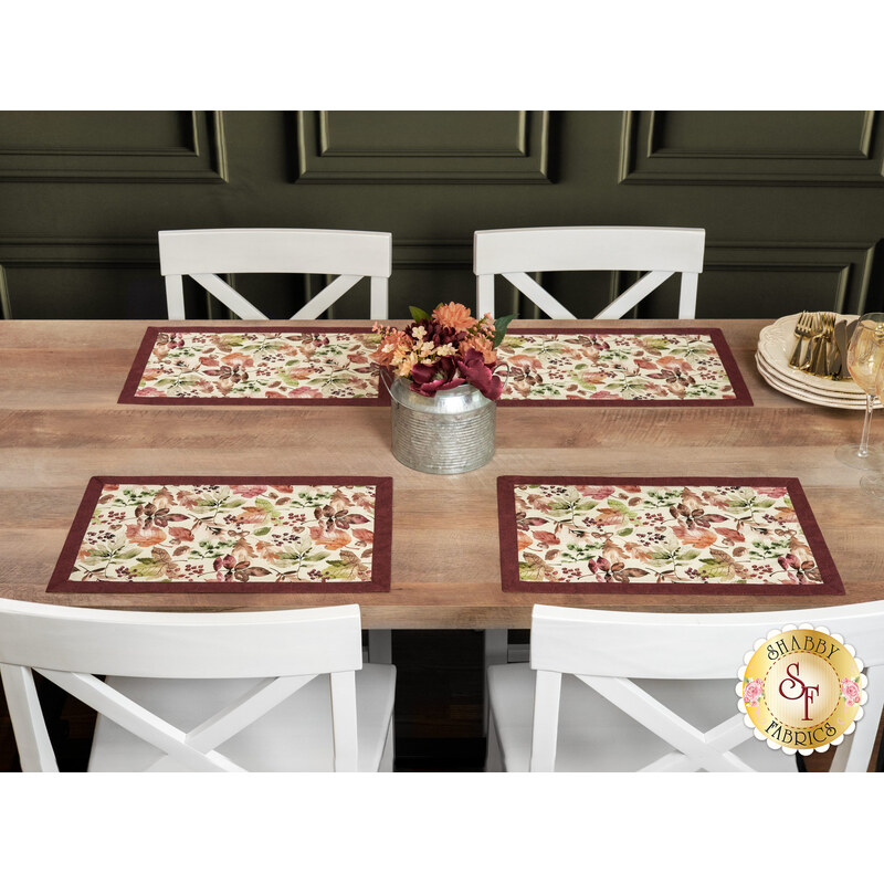 The four completed Self-Binding Placemats, colored in warm autumn fabrics from the Rustle collection by Maywood Studio, staged on a rustic wooden table with white chairs; a tin of coordinating flowers sits in the center.