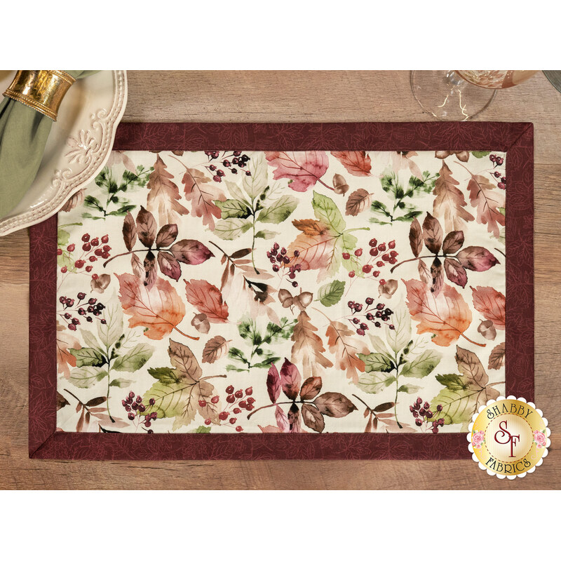 A top down shot of one of the placemats, demonstrating details on the cream fabric with a multicolor leaf print and the wine red fabric used for the binding.