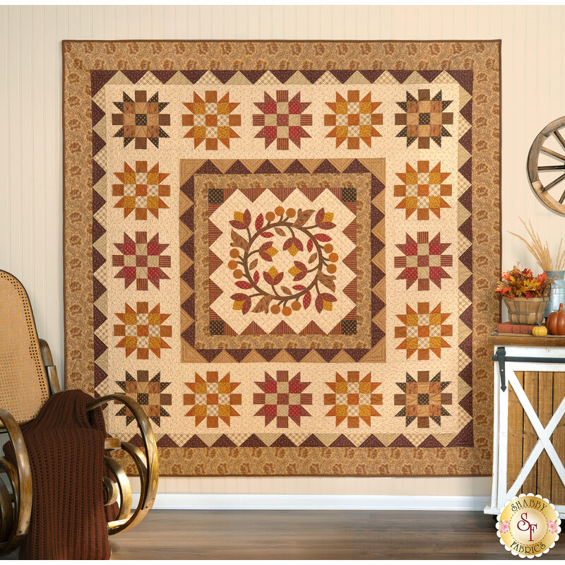 The completed Heartland quilt, a blended design of appliqué and patchwork colored in warm fabrics from the Toasted and Spice Market collections from Andover Fabrics. The project is hung on a light cream wall and staged with rustic farmhouse decor including a rocking chair, coordinating leaves, and a wagon wheel.