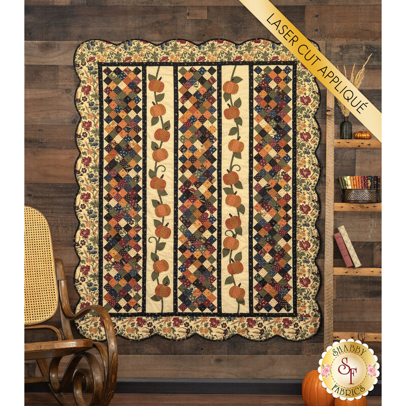 The completed Pumpkin Lane Quilt colored in rich orange, brown, blue, tan, and red fabrics from the Wheatland collection by Moda Fabrics, hung on a rustic wooden wall and staged with coordinating decor between a wooden ladder and vintage rocking chair. A yellow banner in the upper right corner reads 