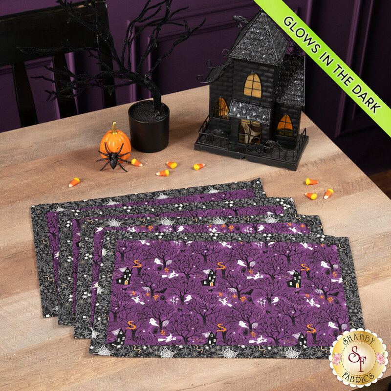 The four completed Self-Binding Placemats, fanned out and colored in purple and gray fabrics from the Witchy Woods collection by Lewis & Irene, staged on a wooden table with coordinating Halloween decor. A lime green banner in the top corner reads 