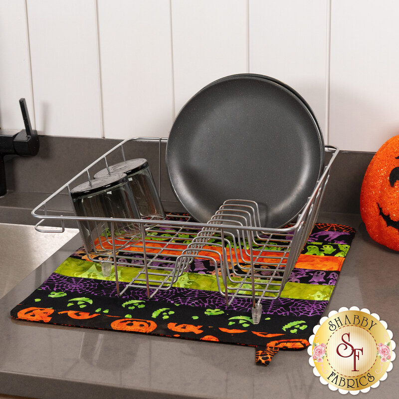 The completed Dry Ideas dish drying mat colored in black, green, purple, and orange fabrics from the Tonga Haunted Batiks collection, staged with a silver dish rack holding a black plate and two glasses 