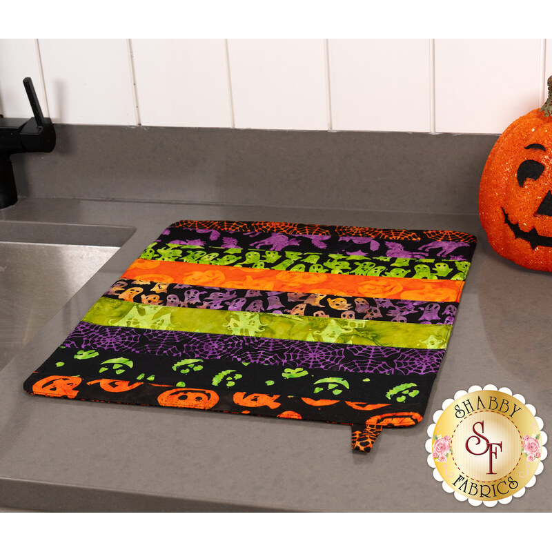 The completed Dry Ideas dish drying mat colored in black, green, purple, and orange fabrics from the Tonga Haunted Batiks collection, staged on a kitchen countertop beside a decorative jack-o'-lantern.
