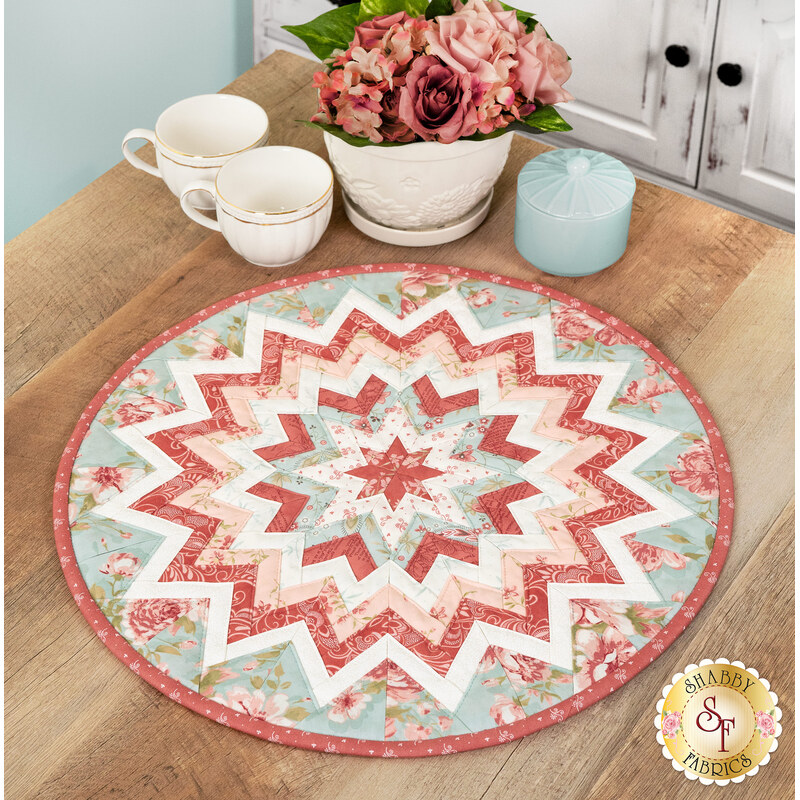The completed Folded Star Table Topper, colored in pink, blue, and white fabrics from the Muse collection, staged on a wooden table with coordinating teacups and flowers.