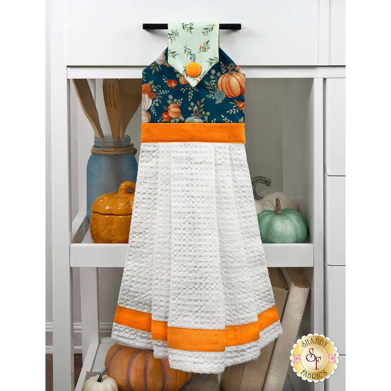 The Hanging Towel, colored in aqua, blue, and orange fabrics from the I Love Fall Most of All collection, staged on a white kitchen drawer with coordinating decor.