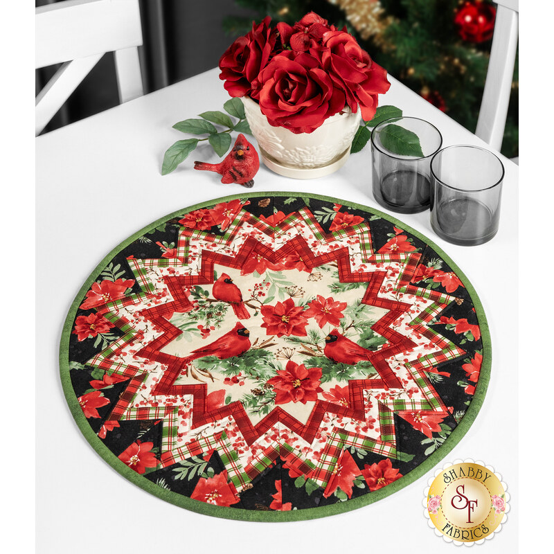 The completed Point of View Folded Star Table Topper colored in black, red, and green fabrics from the Joyful collection. The topper is staged on a white table with coordinating rose decor and tableware.