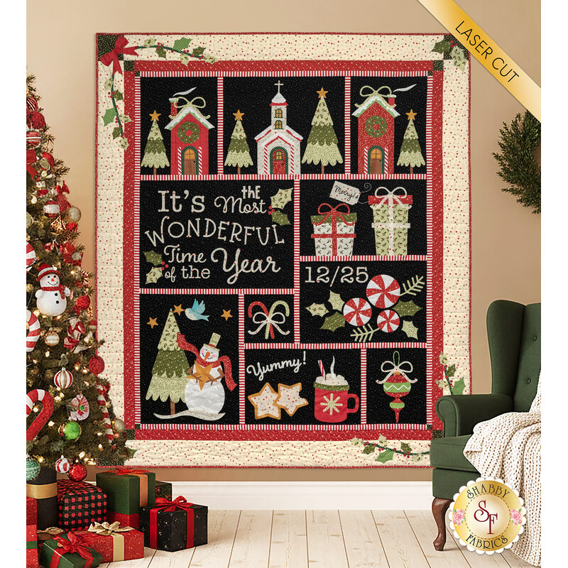 The completed The Most Wonderful Time BOM quilt decorated in red, green, and black Christmas fabrics from the Glisten collection, hung on a tan wall and bordered by cozy Christmas décor like presents, a decorated tree, and a green chair with a blanket thrown over the side.