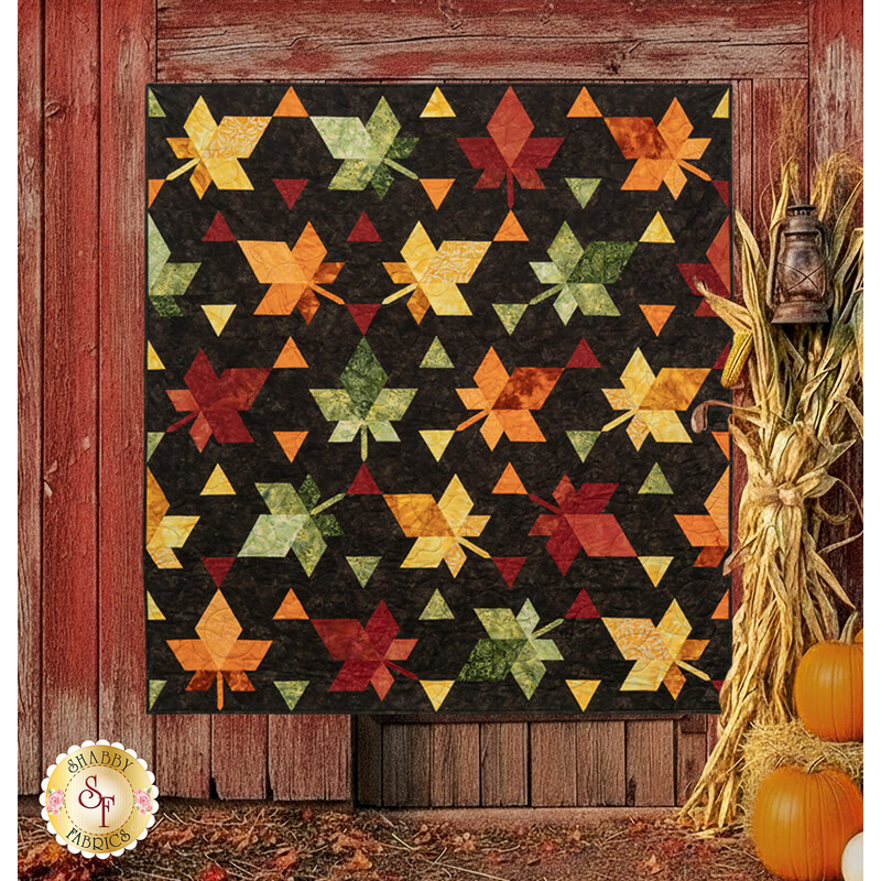 The completed Fall Foliage quilt, colored in contrasting red, orange, green, yellow, and black batik fabrics from the Forest Flora collection, staged on a rustic red barn with pumpkins and cornstalks. 