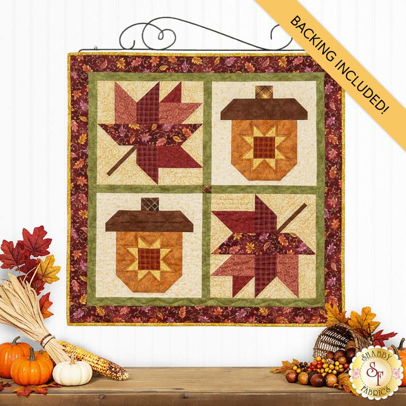 The completed Pieceful Patchwork - November wall hanging, a square project with two patchwork acorns and leaves in autumn themed fabrics, hung on a white paneled wall and staged with coordinating fall decor like corn, pumpkins, and leaves.