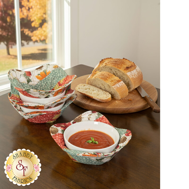 The completed Bowl Cozies, colored in soft autumn fabrics from the Harvest Blooms collection by Wilmington prints, staged on a dark wooden table. Freshly sliced bread sits on a cutting board beside a bowl cozy with a bowl of tomato soup in it and a window on the left side shows an autumn scene outside.