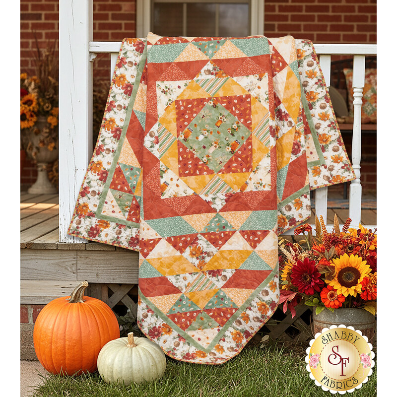 The completed Harvest Blooms Quilt colored in autumn-themed prints from the Harvest Blooms collection by Wilmington prints, artfully draped over a white porch and staged with coordinating fall flowers and pumpkins.