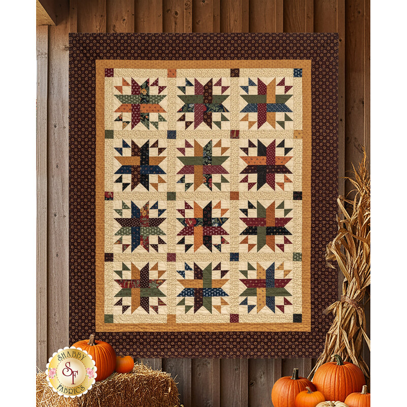 The completed Woven Dreams quilt colored in warm brown, gold, and cream primitive fabrics from the Wheatland collection by Moda Fabrics, hung on a wooden wall and staged with pumpkins and autumnal décor.