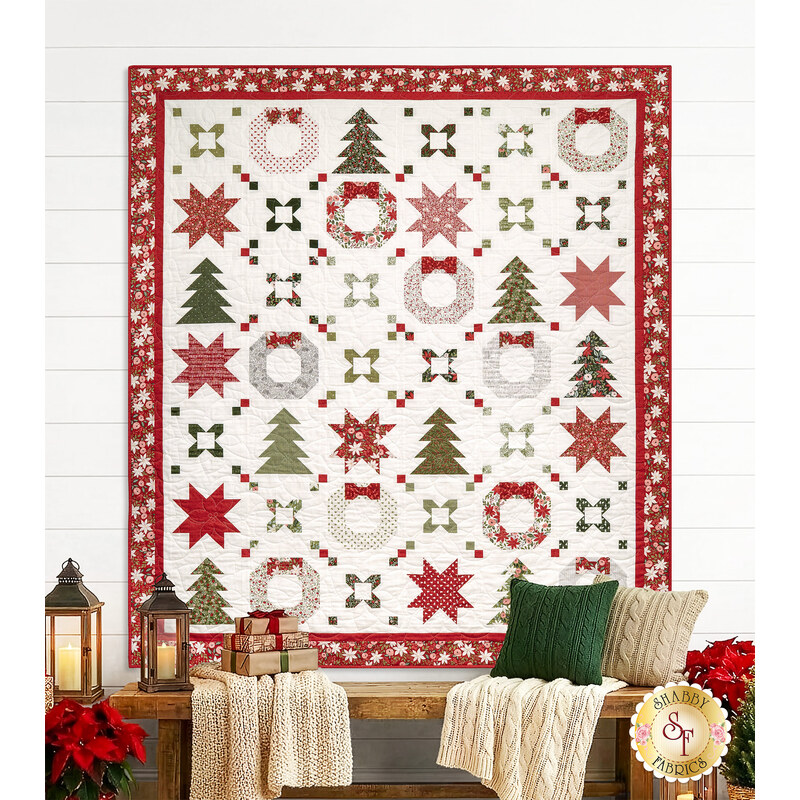 The completed Joyful Connections quilt colored in white, green, and red fabrics from the Berry & Pine collection by Moda Fabrics, hung on a white shiplap wall, staged above a wooden bench with coordinating pillows and blankets.