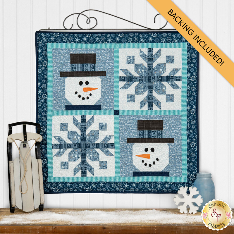 The completed Pieceful Patchwork - January, colored in wintry blue fabrics depicting two patchwork snowmen with appliqué faces and two patchwork snowflake. The project is hung on a white paneled wall and staged with coordinating décor. A yellow banner at the top reads "Backing Included."