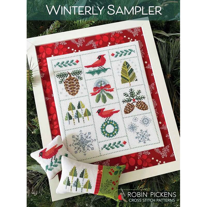 A framed winter-themed cross-stitch sampler with red accents, featuring cardinals, pinecones, and snowflakes.