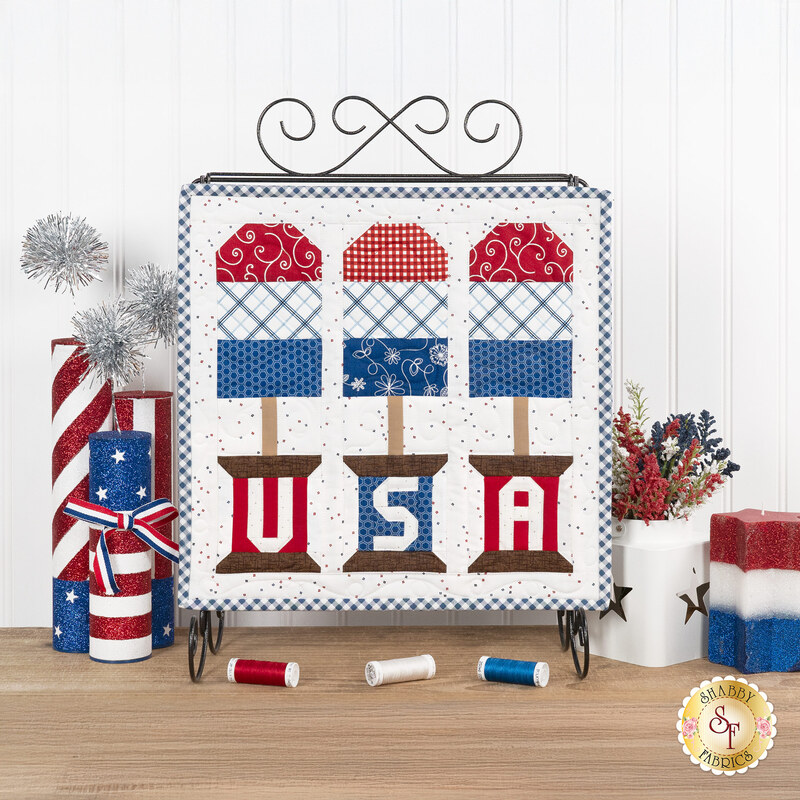 The completed July spool, three popsicles in patriotic colors, hung on a craft holder and staged on a wooden table with coordinating spools and decor.