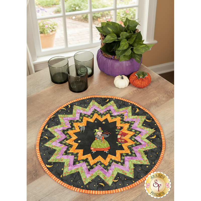 The completed Point of View Folded Star Table Topper colored in black, green, purple, and orange fabrics from the Ghoul Friends collection. The topper is staged on a wood table with coordinating Halloween decor and tableware.