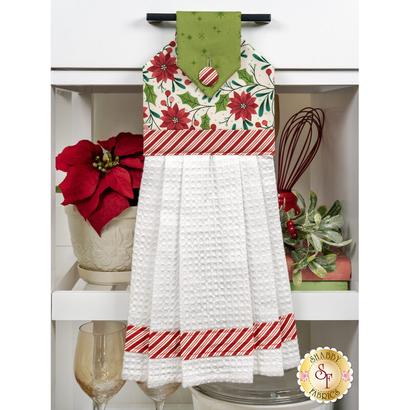 The completed hanging towel staged on a kitchen drawer, colored in white, green, and red fabrics from the Make it Merry collection. Coordinating red decor peeks in from behind the towel.