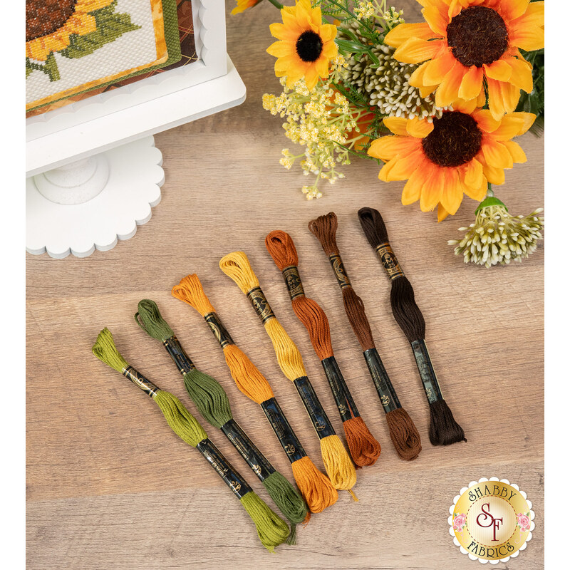 The seven piece embroidery floss set for the Learn to Cross Stitch Sunflower project, staged on a wooden table beside the finished sunflower project and coordinating decorative flowers.
