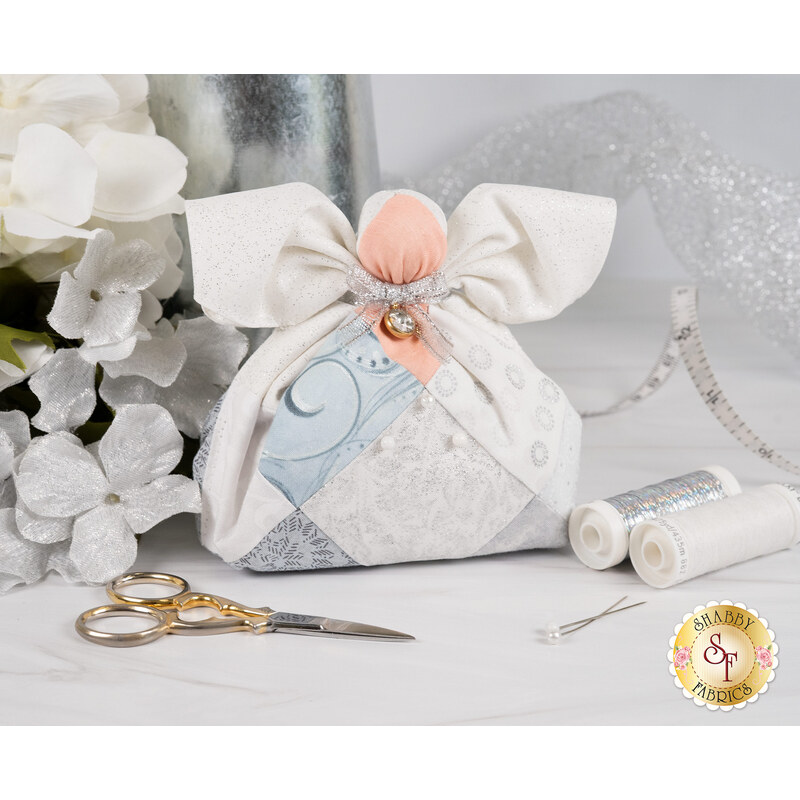 The completed April Birthstone Angel with a silver and white palette, white wings, a silver bow and silver charm beside coordinating decor and notions.