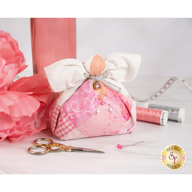 The completed October Birthstone Angel with a rosy palette, white wings, and a silver bow and blush pink charm beside coordinating decor and notions.