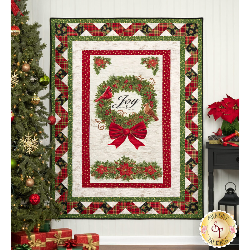 The completed Ribbon Border Panel Quilt colored in red, green, cream, and gold metallic fabrics from the Joyful collection by Timeless Treasures, hung on a white paneled wall and staged with a decorated tree, poinsettia flowers, and presents in coordinating colors.