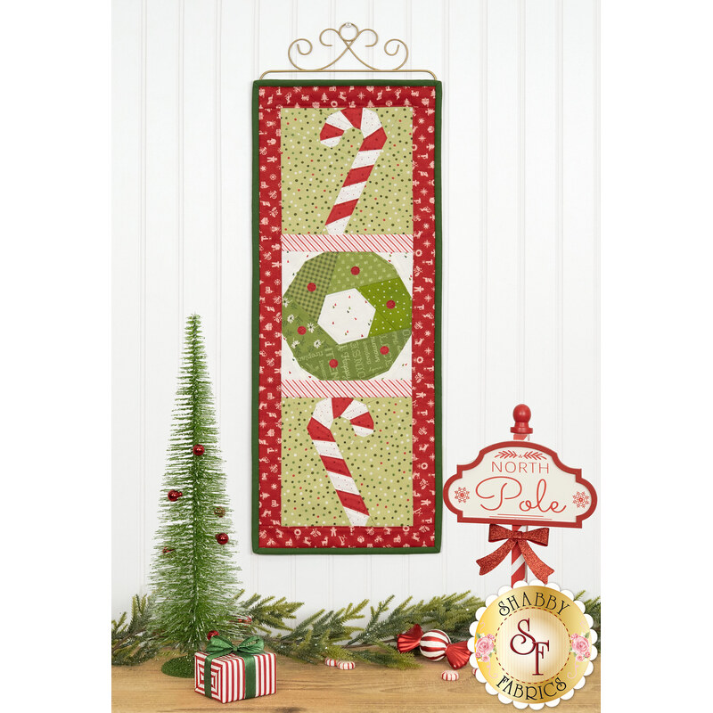 The completed December project, depicting two candy canes and a green wreath, hung on a craft hanger against a white paneled wall and staged with coordinating, playful Christmas decor.