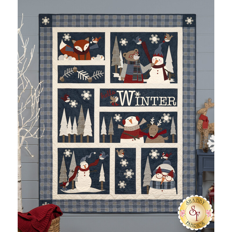 The completed Woodland Snowmies - Making Friends quilt colored in blue, cream, and cool-toned flannel fabrics from the Prairie Gatherings Flannel collection by Moda Fabrics, hung on a blue-paneled wall and surrounded by coordinating winter-themed décor.