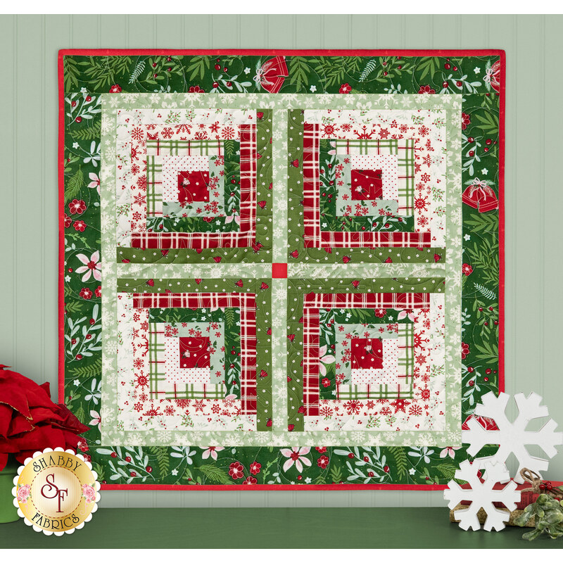 The completed Log Cabin Wall Hanging colored in cream, green, and red Christmas fabrics from the Mistletoe & Holly collection, hung on a mint paneled wall and staged with coordinating Christmas decor.