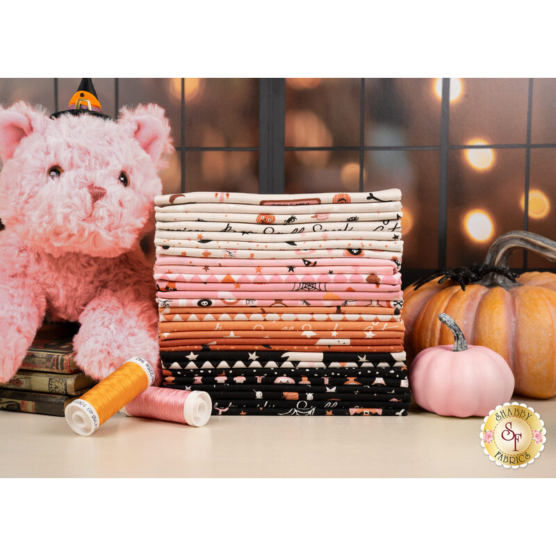 A stack of the fabrics included in the Purrs and Boos FQ set, with a plush cat and books and pumpkins and a spider and thread.