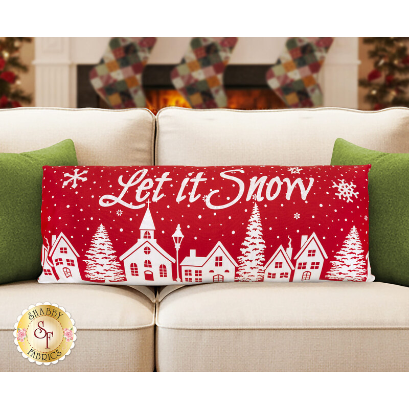 The completed Let it Snow Pillow colored in the festive fabrics from the Buon Natale collection, staged on a neutral couch in front of a decorated Christmas fireplace with stockings.