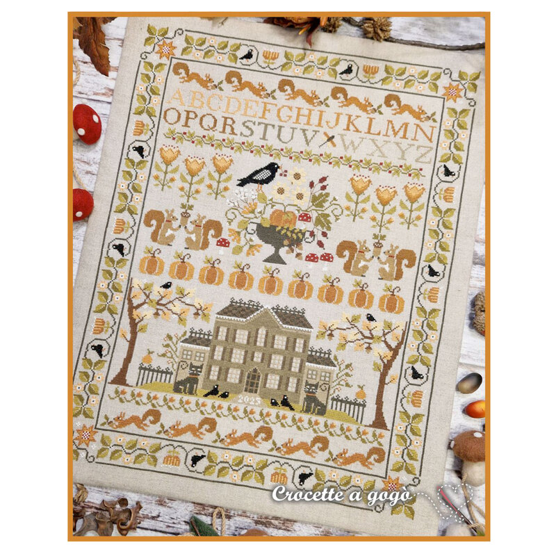 The finished cross-stitch sampler featuring a house, floral motifs, pumpkins, squirrels, and crows surrounded by coordinating autumn decor.  