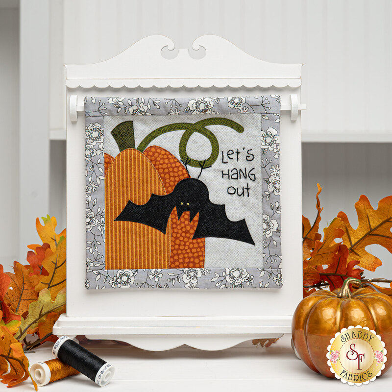 Colorful wall hanging featuring a bat and pumpkins with the text Let's hang out, hung on a white display board.