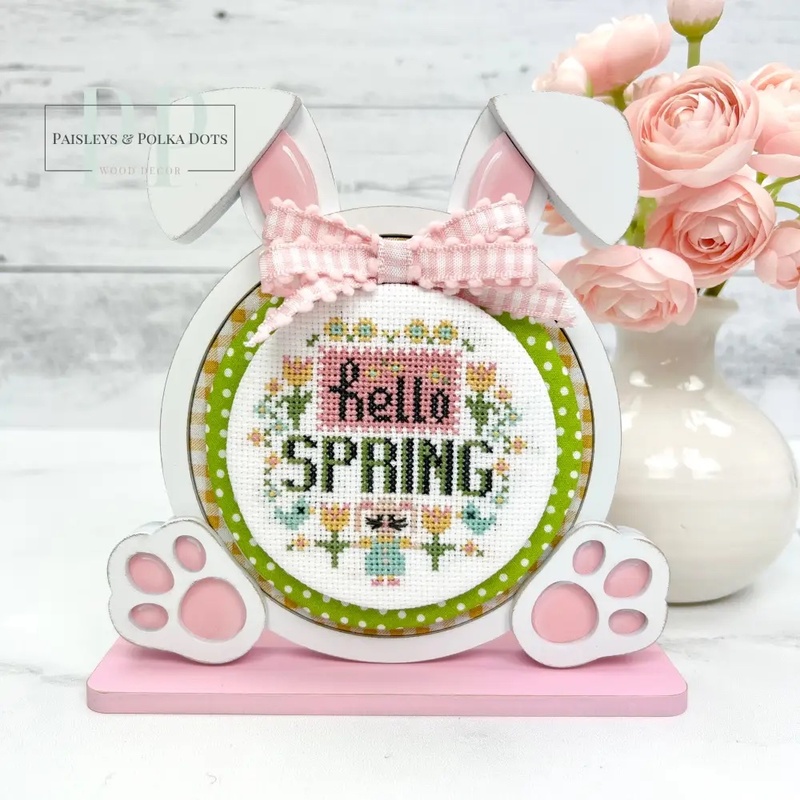 The fully assembled and painted Bunny Round Cross Stitch Display Frame, staged on a white tabletop next to a vase of pink roses, displaying a festive cross-stitch project.