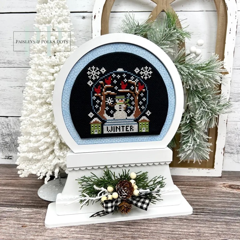The fully assembled and painted Snow Globe Wood Display, staged on a white tabletop next to a tiny decorative tree, displaying a festive cross-stitch project.