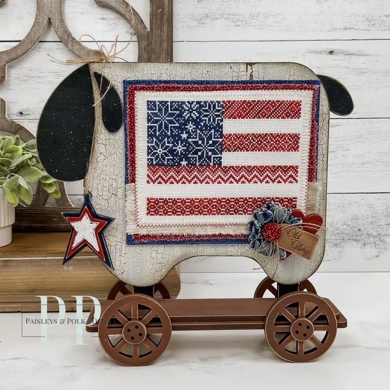 The fully assembled and painted Primitive Sheep Cross Stitch Display, staged on a white tabletop next to a decorative wall shelf, displaying a festive cross-stitch project.