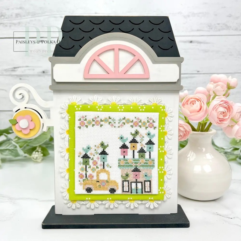 The fully assembled and painted City Flower Shop Cross Stitch Display Backer, staged on a marble countertop with coordinating flowers, displaying a spring cross-stitch project.