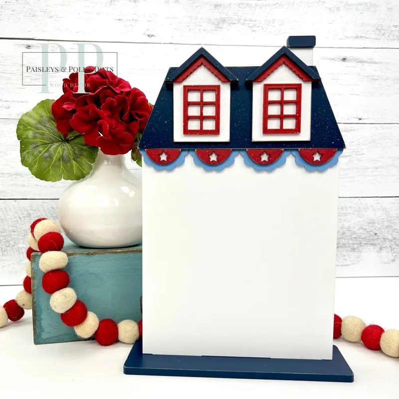 The fully assembled and painted Patriotic City House #3 Cross Stitch Display, staged on a white tabletop with coordinating patriotic décor and flowers.