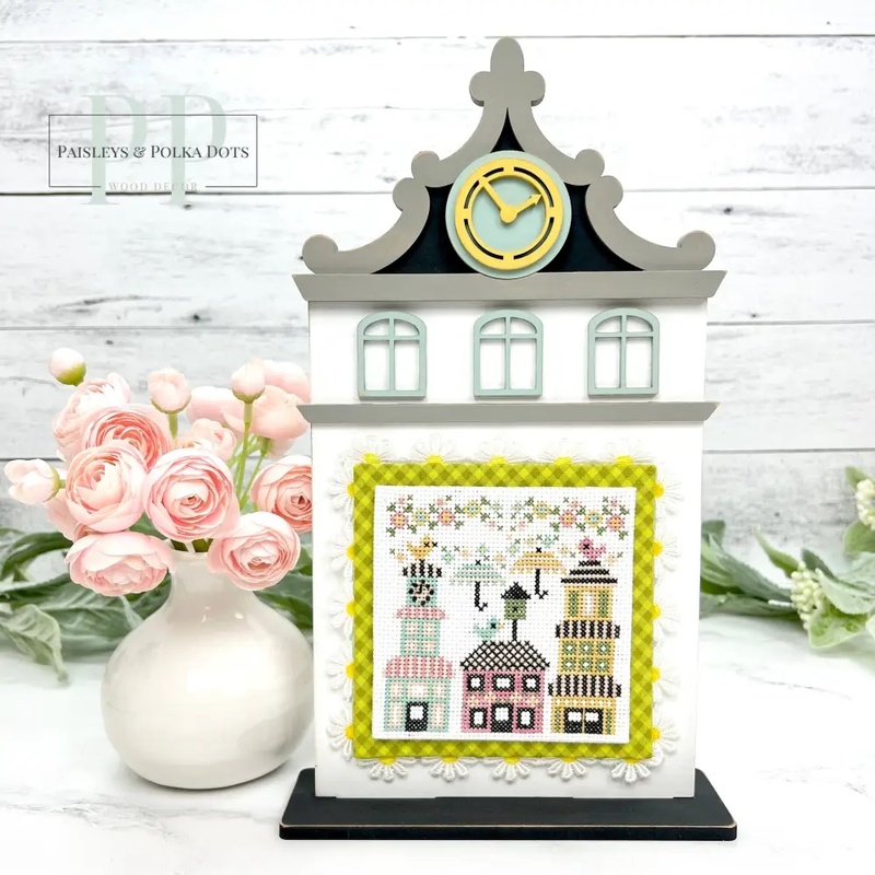 The fully assembled and painted City Clock Tower Cross Stitch Display Backer, staged on a marble countertop with coordinating flowers, displaying a spring cross-stitch project.